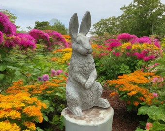 Può includere: Una statua di coniglio in cemento grigio in piedi su un piedistallo in cemento grigio in un giardino con fiori colorati.