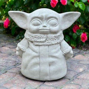 May include: A light gray concrete statue of a character with large ears, a textured scarf, and a long coat. The statue is set on a brick patio with a backdrop of green foliage and vibrant pink flowers.