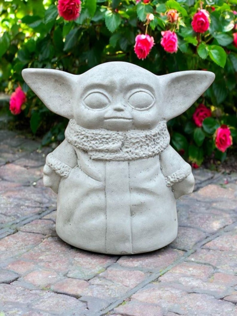 May include: A light gray concrete statue of a character with large ears, a textured scarf, and a long coat. The statue is set on a brick patio with a backdrop of green foliage and vibrant pink flowers.