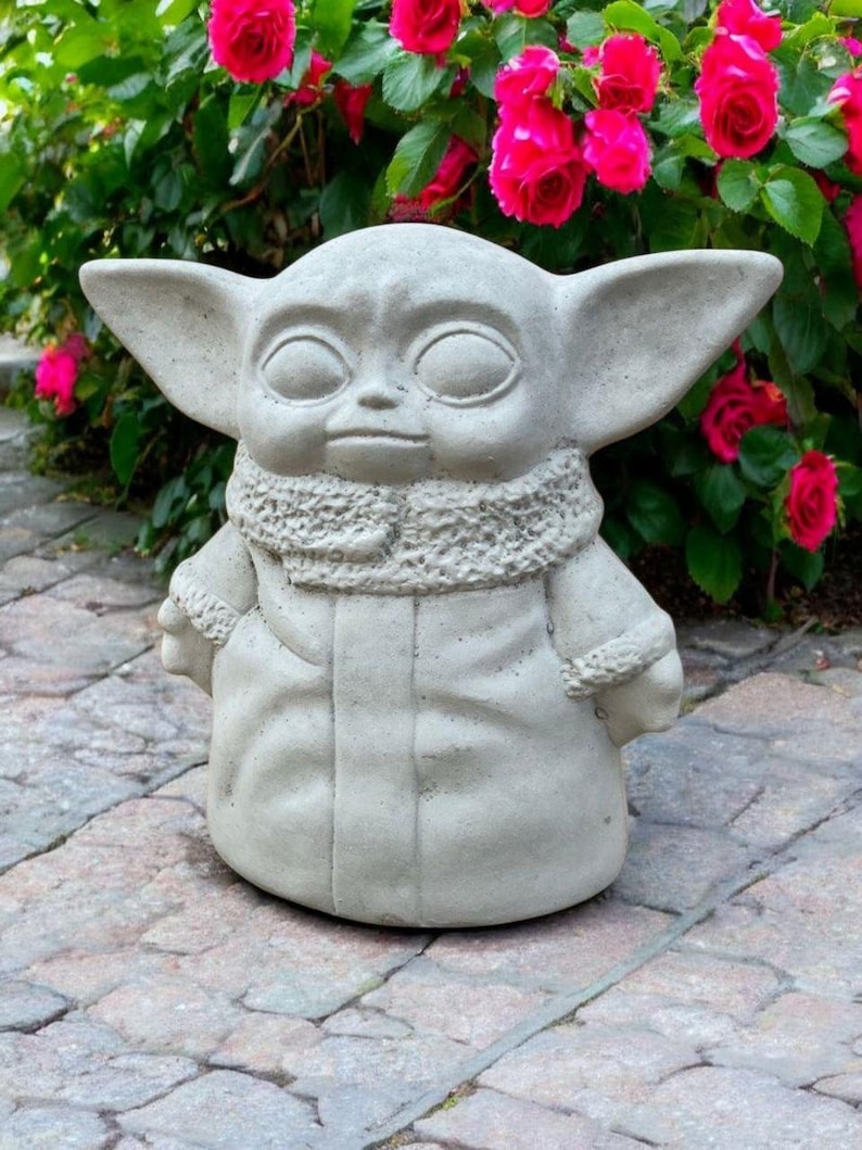 May include: A light gray concrete statue of a character with large ears and a hooded coat. The statue is standing on a stone surface, with vibrant pink roses in the background. The statue has a detailed texture and a serene expression.