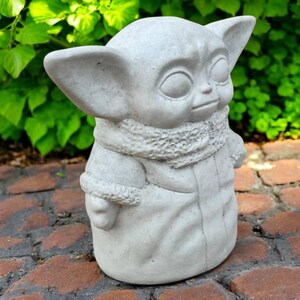 May include: A light gray statue of a character with large ears and a hooded robe. The statue has a textured surface and is standing on a brick surface. The statue is a popular character from a science fiction franchise.