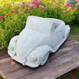 May include: A grey concrete planter shaped like a classic Volkswagen Beetle convertible. The planter has a rounded design with a detailed front grille and headlights.