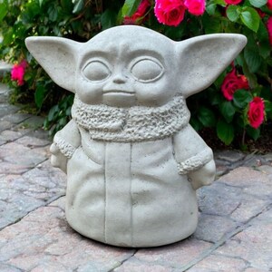 May include: A light gray concrete statue of a character with large ears and a hooded coat. The statue is standing on a stone surface, with vibrant pink roses in the background.