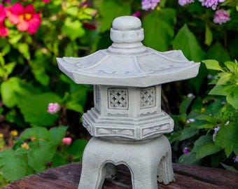 May include: A gray stone Japanese garden lantern with a multi-tiered design. The lantern has a pagoda-style roof and decorative cutouts. The lantern is set on a wooden surface, with colorful flowers in the background.