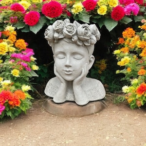 May include: A white concrete sculpture of a girl's head with closed eyes. The sculpture is wearing a wreath of roses and has a detailed floral pattern on the chest. The sculpture is sitting on a brown base.