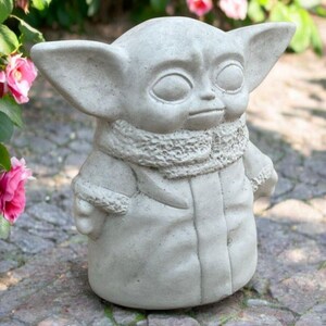 May include: A gray concrete statue of a character with large ears, large eyes, and a textured scarf. The statue is wearing a coat with a textured collar. The statue is standing on a stone surface, with pink flowers and green foliage in the background.
