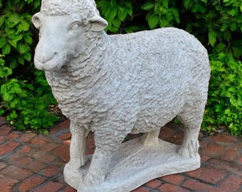 May include: A light gray concrete sheep statue with detailed wool texture. The sheep stands on a rectangular base, with a green leafy background. This outdoor garden decor adds a touch of rustic charm.