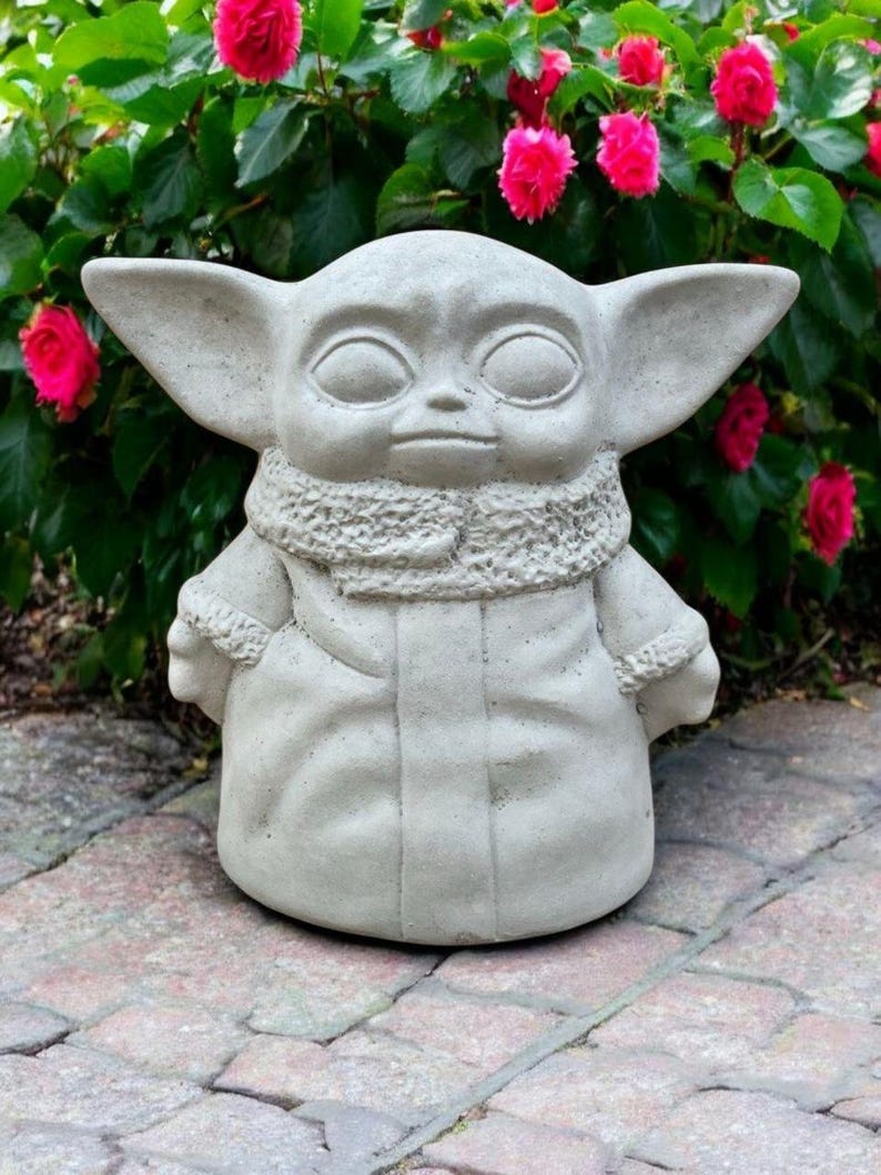 May include: A light gray concrete statue of a character with large ears, a large head, and a robe. The statue is outdoors, in front of a green bush with bright pink flowers. The statue is sitting on a brick surface.