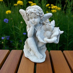 May include: A small, gray stone angel statue. The angel is seated, with wings and a thoughtful expression, holding a flower. The statue is on a wooden surface, with a background of yellow flowers and green foliage.