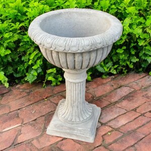May include: A gray, ornate stone planter with a wide, scalloped rim and a fluted pedestal base. The planter sits on a square base and is surrounded by green foliage and red brick pavers.