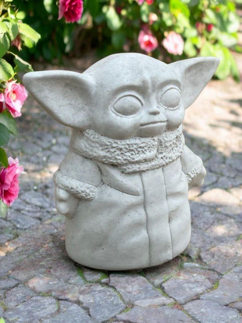 May include: A gray concrete statue of a character with large ears and a hooded coat. The statue has a detailed texture and is set outdoors with pink flowers in the background. The figure is a popular character from a science fiction series.