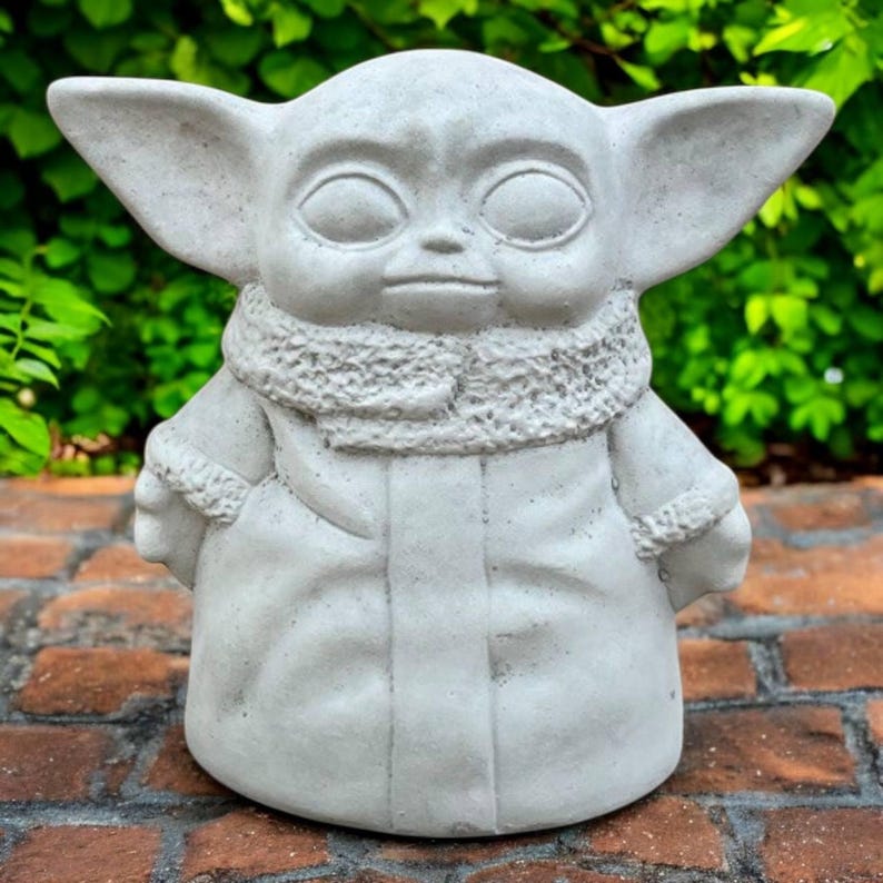 May include: A gray concrete statue of a character with large ears, large eyes, and a textured scarf. The figure is wearing a coat and has its hands clasped in front. The statue is on a brick surface with green foliage in the background.