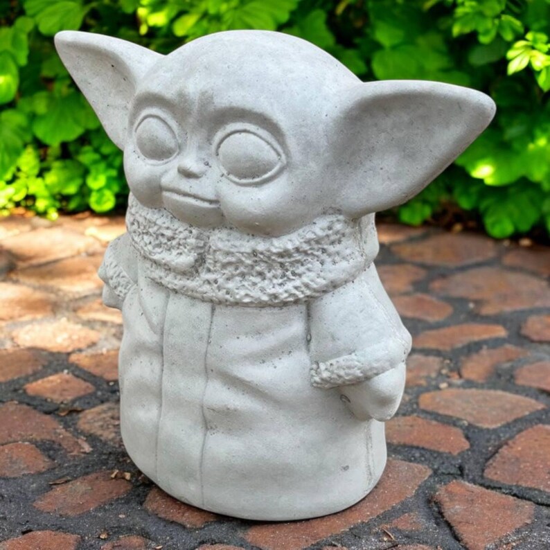 May include: A light gray concrete statue of a character with large ears, large eyes, and a small mouth. The statue is wearing a coat with a fluffy collar. The statue is standing on a brick surface with green foliage in the background.