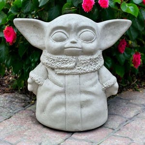 May include: A light gray concrete statue of a character with large ears, a large head, and a robe. The statue is outdoors, in front of a green bush with bright pink flowers. The statue is sitting on a brick surface.
