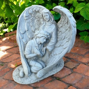 May include: A grey stone angel statue with large, detailed wings. The angel embraces a kneeling figure, both with serene expressions. The statue is set on a brick surface, with green foliage in the background.