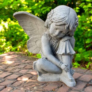 May include: A gray concrete fairy statue with large, detailed wings. The fairy is seated, with its head bowed and arms wrapped around its legs. The statue is on a brick surface, with a blurred green background.