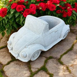 May include: A light grey concrete planter in the shape of a classic convertible car. The planter has a large open space for plants and sits on a stone surface. Red flowers are in the background.