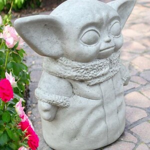 May include: A light gray concrete statue of a character with large ears and a hooded coat. The statue has a detailed scarf and is standing on a brick surface. Pink and red roses are in the background.