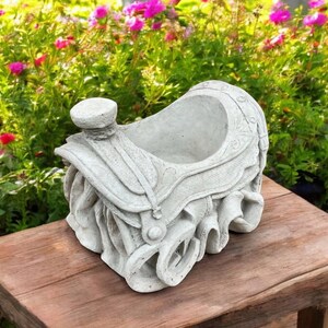 May include: A white concrete planter shaped like a saddle, with a saddle horn and stirrups. The planter is sitting on a wooden table.