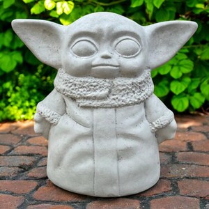 May include: A light gray statue of a character with large ears, large eyes, and a textured collar. The statue is wearing a robe and has a neutral expression. The statue is sitting on a brick surface with green foliage in the background.