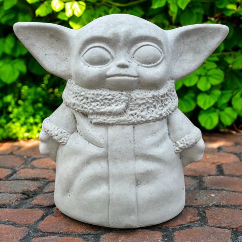 May include: A light gray statue of a character with large ears, large eyes, and a textured collar. The statue is wearing a robe and has a neutral expression. The statue is sitting on a brick surface with green foliage in the background.