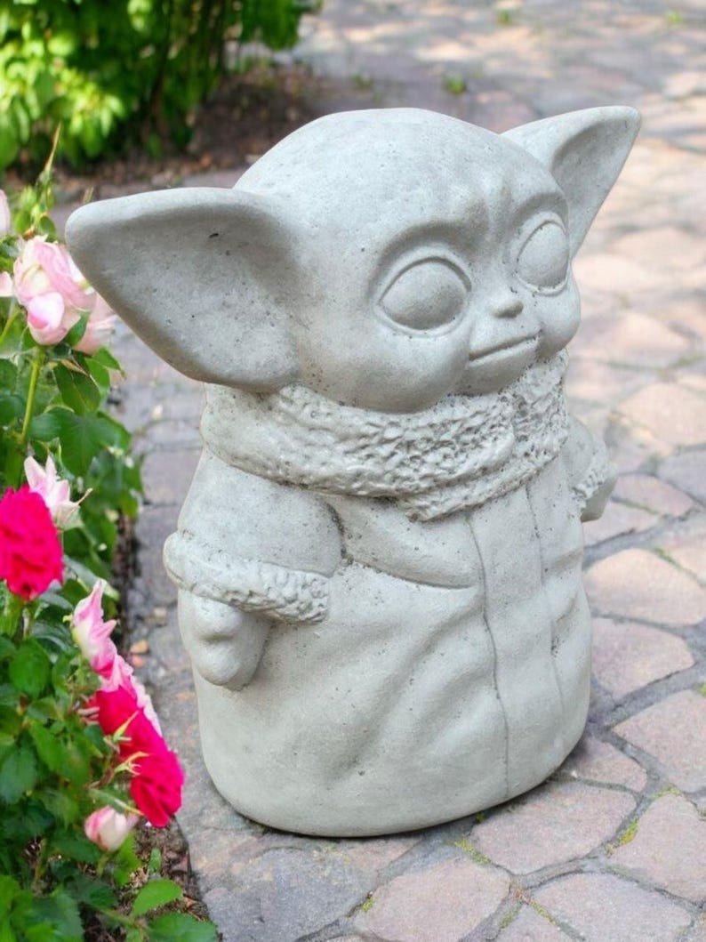 May include: A light gray concrete statue of a character with large ears and a hooded coat. The statue has a detailed scarf and is standing on a brick surface. Pink and red roses are in the background.