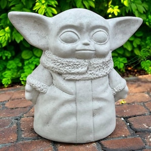 May include: A light gray concrete statue of a character with large ears, large eyes, and a textured scarf. The statue is wearing a coat and has its hands clasped in front. The statue is set on a brick surface with green foliage in the background.