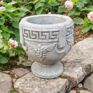 May include: A gray stone planter with a wide, circular opening and a pedestal base. The exterior features a relief pattern of a Greek key design around the upper rim and a cluster of grapes with leaves carved into the center of the bowl.