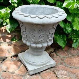 May include: A gray, ornate outdoor planter with a scalloped rim and leaf pattern. The planter sits on a square base and is made of a stone-like material. The background features green foliage and a brick surface.