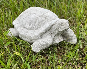 May include: A gray concrete turtle statue with a detailed shell and head. The turtle is positioned on its belly with its legs outstretched.