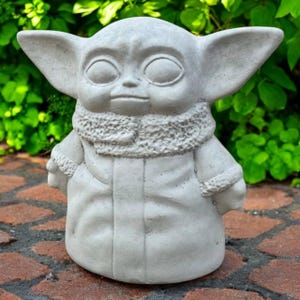 May include: A light gray statue of a character with large ears and large eyes, wearing a coat with a fur-like collar. The statue is standing on a brick surface with green foliage in the background.