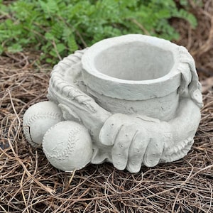 May include: A gray concrete planter shaped like a baseball glove holding two baseballs. The planter has a round opening for planting.