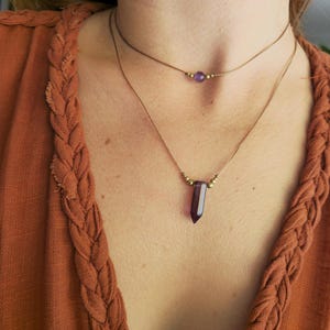 Könnte beinhalten: Zwei feine Halsketten. Die obere Halskette hat eine kleine lila Perle mit goldenen Details. Die untere Halskette hat einen dunkelvioletten Kristallanhänger mit goldenen Akzenten. Die Halsketten werden auf einem rostfarbenen Oberteil getragen.