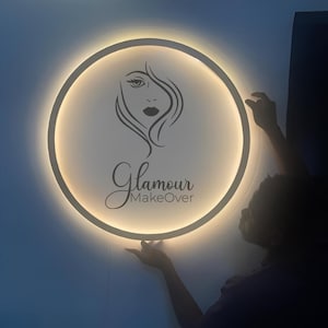 May include: A circular wall sign featuring a backlit white surface with a black line art illustration of a face and the text Glamour MakeOver. The sign emits a soft warm glow from behind its circular frame, creating a halo effect.