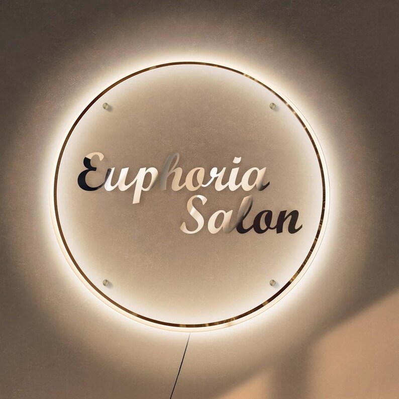 May include: Circular illuminated sign with the text "Euphoria Salon" in a stylish font. The sign has a gold-colored trim and is mounted on a neutral-toned wall. The sign is lit with a warm, inviting glow.