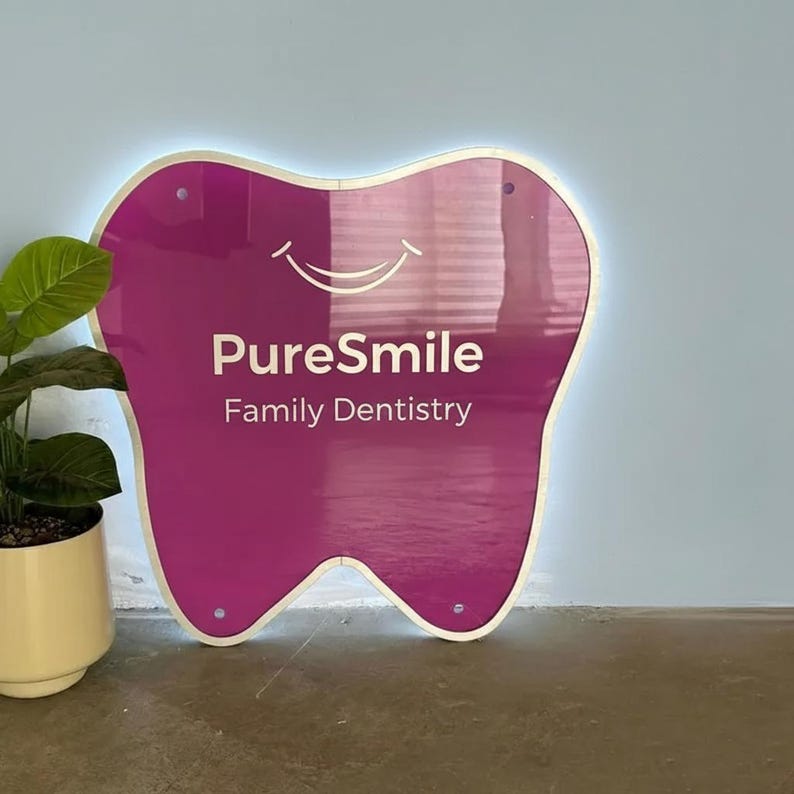 Op de afbeelding: Een magenta tandvormig bord met de tekst "PureSmile Family Dentistry" in het wit. Het bord heeft een witte rand met een zachte gloed en is bevestigd aan een lichtblauwe muur. Links is een plant in een pot te zien.