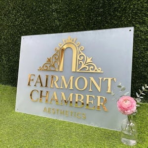 May include: A rectangular frosted acrylic sign featuring gold-colored raised lettering and a decorative arch emblem. The text reads FAIRMONT CHAMBER AESTHETICS. The sign is positioned upright with a single pink flower in a glass vase nearby.