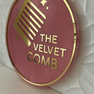 May include: Round sign with a pink velvet background and gold trim. The sign features a gold comb graphic, gold stars, and the text "THE VELVET COMB" in gold lettering. The sign is mounted on a white wall.