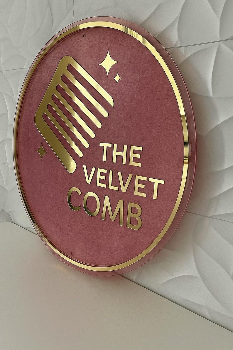 May include: Round sign with a pink velvet background and gold trim. The sign features a gold comb graphic, gold stars, and the text "THE VELVET COMB" in gold lettering. The sign is mounted on a white wall.