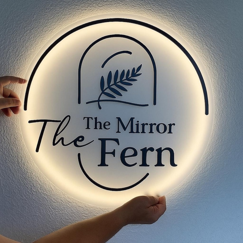 Op de afbeelding: Een ronde, verlichte sign met de tekst "The Mirror Fern" in een stijlvol lettertype. Het bord heeft een zwarte rand en een centraal ontwerp van een varen in een boog. Het bord wordt door twee handen vastgehouden.