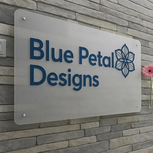May include: A rectangular sign with the text "Blue Petal Designs" in blue, along with a floral graphic. The sign is mounted on a gray brick wall. A pink gerbera daisy is in a clear vase to the right.