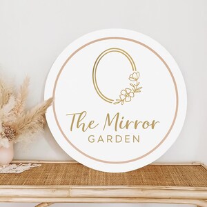 May include: A round white sign with a gold floral design and the text "The Mirror Garden" in gold script. The sign has a light pink border and is displayed on a woven table.