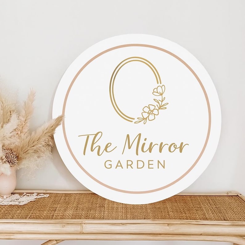 May include: A round white sign with a gold floral design and the text "The Mirror Garden" in gold script. The sign has a light pink border and is displayed on a woven table.