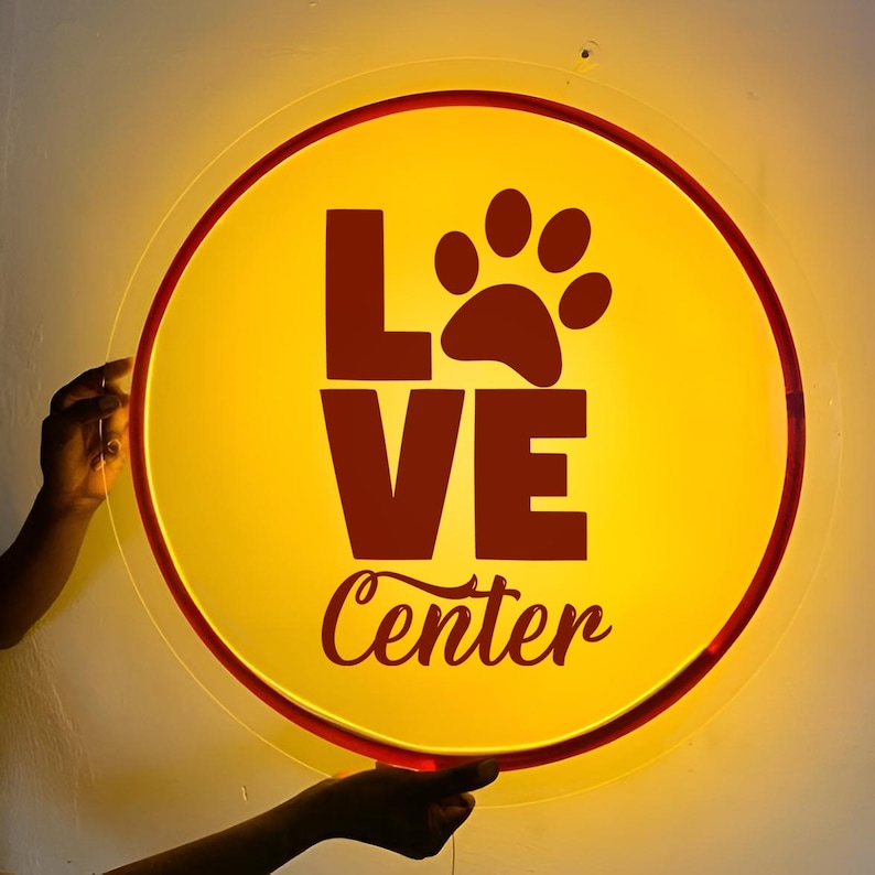 May include: A circular, illuminated sign with a bright yellow background and a dark red border. The sign features the word LOVE in large, bold, dark red letters, with a paw print replacing the letter O. Below, the word Center is written in a dark red script font.