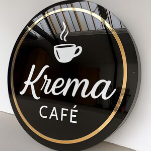 May include: Round black sign with gold trim, featuring the white text "Krema Caf&eacute;" and a white coffee cup icon with steam. The sign is mounted on a white wall, suggesting a cafe or coffee shop.