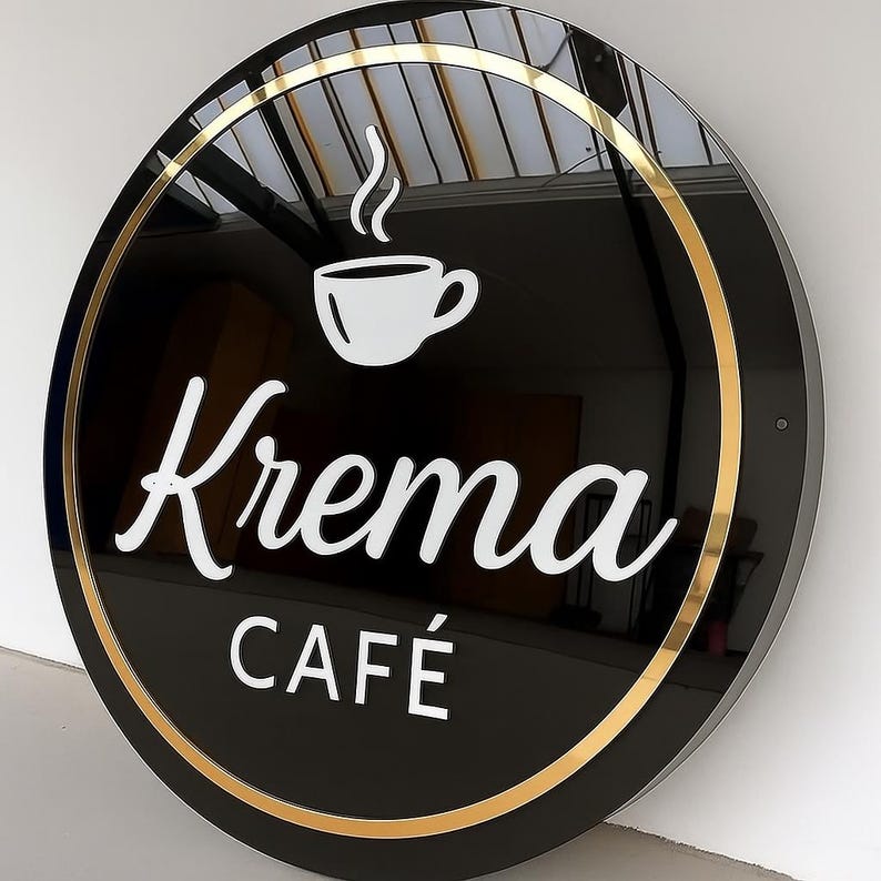 May include: Round black sign with gold trim, featuring the white text "Krema Caf&eacute;" and a white coffee cup icon with steam. The sign is mounted on a white wall, suggesting a cafe or coffee shop.