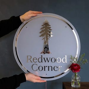 May include: A circular sign with a frosted white surface and a reflective silver border. The sign features a metallic silver tree graphic centered above the text "Redwood Corner" in a serif font. The design includes small mounting holes.