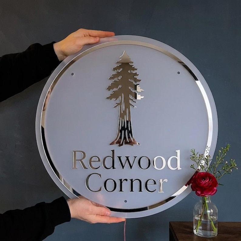May include: A circular sign with a frosted white surface and a reflective silver border. The sign features a metallic silver tree graphic centered above the text "Redwood Corner" in a serif font. The design includes small mounting holes.