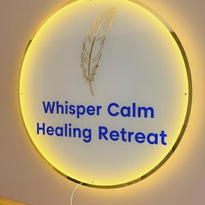 Op de afbeelding: Een ronde, verlichte bewegwijzering met een gouden rand en een gouden veergrafiek. Op het bord staat "Whisper Calm Healing Retreat" in blauwe tekst. Het bord is op een lichtbruine muur gemonteerd.