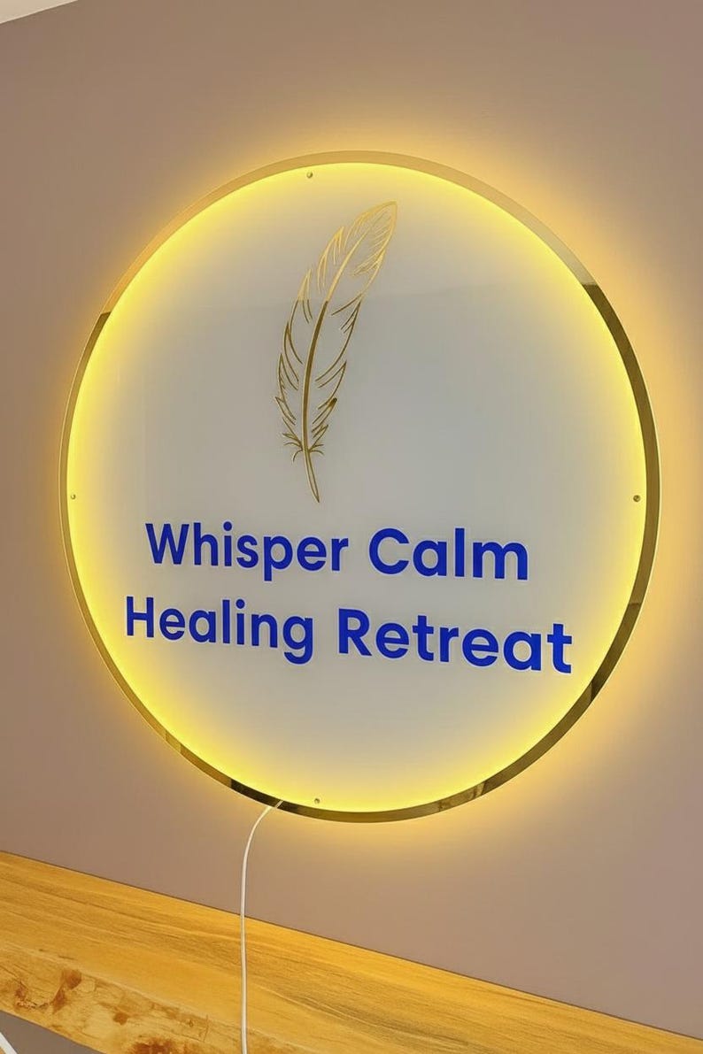 Op de afbeelding: Een ronde, verlichte bewegwijzering met een gouden rand en een gouden veergrafiek. Op het bord staat "Whisper Calm Healing Retreat" in blauwe tekst. Het bord is op een lichtbruine muur gemonteerd.